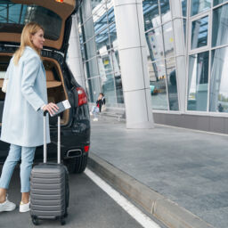 Back view of woman with boarding ticket and trolley suitcase standing at rear of vehicle